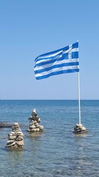 Greek flag waving in wind with balanced stones by the sea