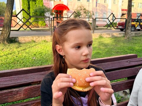 Children and fast food. A cute little girl eats a hamburger on a bench in an outdoor park.