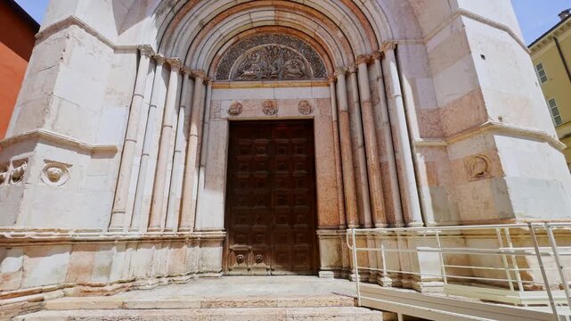 Front view of the Baptistery of Parma entrance, showing its carved Romanesque marble portal, wooden doors, sculpted tympanum and accessibility ramp in Parma, Italy.