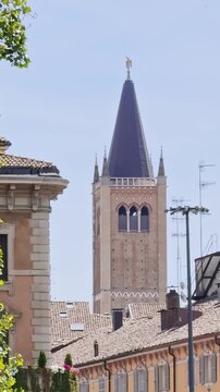 Vertical telephoto view of Parma Cathedral&rsquo;s Gothic bell tower rising above terracotta rooftops, with its steep octagonal spire and gilded angel weather vane against a blue sky.