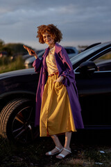 Fototapeta premium Young woman with curly hair walks beside a dark car, wearing a purple coat, striped top, and yellow skirt, expressing a vibrant outdoor lifestyle and confident casual fashion.
