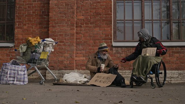 Senior Caucasian homeless man sitting on street sidewalk sharing food with Caucasian with disability in wheelchair. Ideal for social awareness campaigns and charity editorial projects