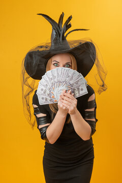 Young woman in a witch hat and black dress holds a fan of dollar bills over her face, eyes peeking out with playful, mysterious expression against a yellow background