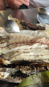 Vertical 4K video showing hands using spoon and fork to peel back charred salt crust skin of grilled snakehead fish to reveal white meat.