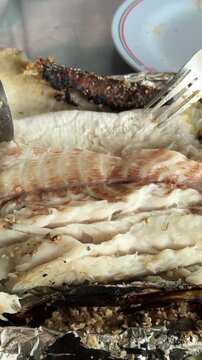 Vertical 4K close up video of hands using cutlery to separate fillet meat and remove bone from salt-crusted grilled snakehead fish.