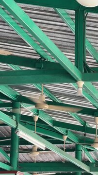 Vertical 4K video showing multiple ceiling fans spinning under green steel roof truss structure in an open air cafeteria or warehouse building.