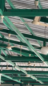 Vertical 4K video showing multiple ceiling fans spinning under green steel roof truss structure in an open air cafeteria or warehouse building.