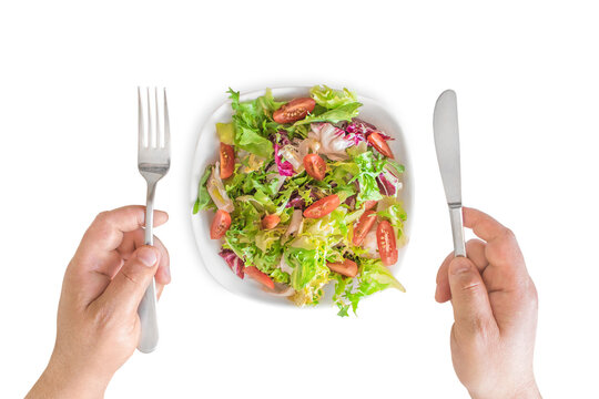 A man's hands holding cutlery next to a white plate containing a healthy salad (cherry tomatoes, mixed greens, arugula, iceberg lettuce) on a white background; top view. Healthy eating and diet concep