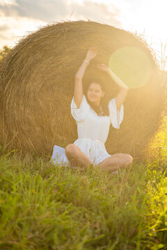 Young woman in white dress relaxing beside a hay bale in warm sunset light. Peaceful countryside moment represents slow living, solitude and connection with nature