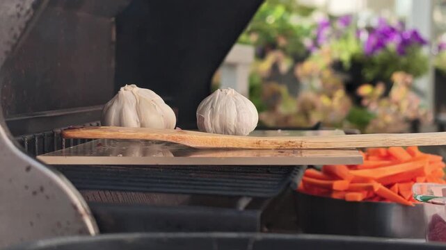 Two whole garlic bulbs sit side by side on worn wooden cutting board placed across metal grill surface beneath open grill hood, with garlic, bulb, wood, cutting detail, showing board, grill, metal