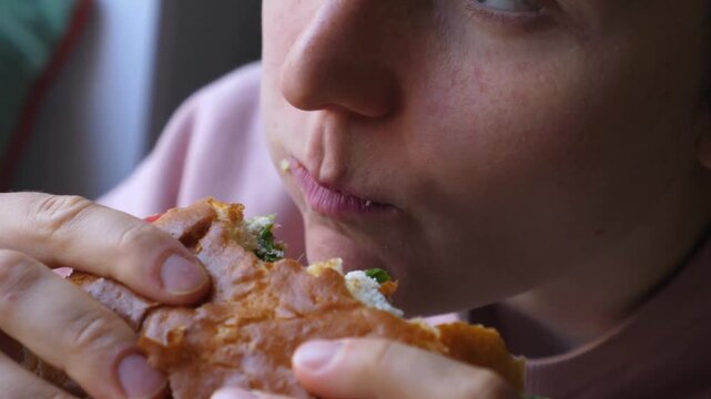 Close up of young woman eating sandwich with relaxed expression. Woman takes repeated bites of crusty bread, chewing filling during simple meal and showing peaceful lunch moment indoors.