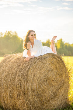 Young woman in white dress lying barefoot on hay bale at sunset. Carefree summer moment represents slow living, relaxation and freedom in countryside