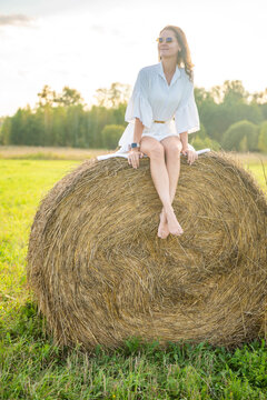 Young woman in white dress sitting barefoot on hay bale in summer countryside. Peaceful rural lifestyle represents slow living, freedom and relaxation in nature