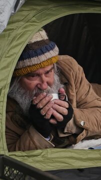 Vertical shot of senior Caucasian homeless man drinking hot beverage from metal mug inside tent. Clip ideal for non-profit charity campaigns, social welfare awareness, and documentary storytelling