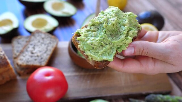 Hand holds avocado toast above cutting board during food prep. Fresh bread is topped with mashed avocado near tomatoes and halved avocados, creating bright healthy snack moment in kitchen light.