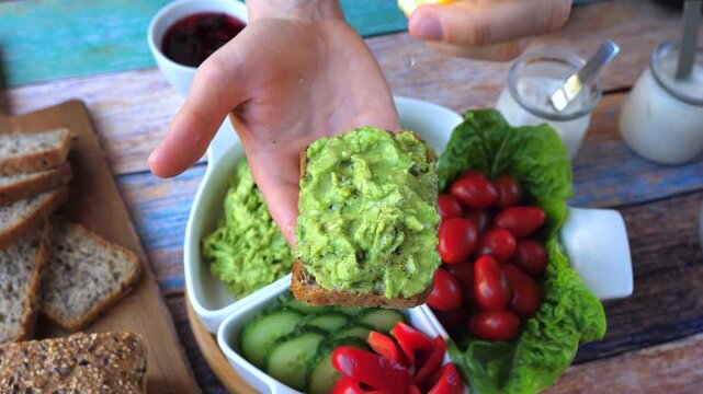 Hands drizzle lemon over avocado toast, finishing colorful breakfast spread. Rustic tabletop holds sliced bread, cucumbers, cherry tomatoes and greens in fresh, inviting food preparation moment.