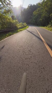 POV cycling footage on a winding mountain road surrounded by dense forest, warm sunlight and long shadows, with a guardrail and mountain scenery in the background.