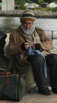 Vertical video of senior Caucasian homeless man sitting outdoors and mending garments. Visuals suit non-profit charity campaigns, social welfare promos, and poverty documentaries