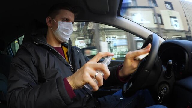 Driver wearing mask sanitizes car interior while traveling through city streets. He sprays and wipes steering wheel with deliberate attention, combining hygiene routine with tense concentration.