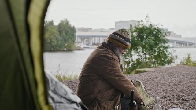 Senior Caucasian homeless man sorting glass bottles into bag near tent outdoors. Footage suitable for social issues documentaries, poverty awareness campaigns, and charity video projects