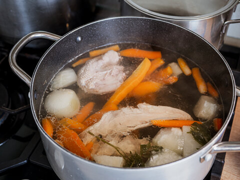 Homemade chicken broth cooking in a large pot with chicken meat, carrots, onion and root vegetables, showing traditional soup preparation on a kitchen stove.