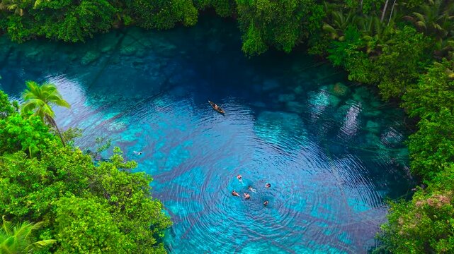 Aerial view of a man and a woman rowing a small canoe and swimming in a clear blue lake surrounded by tropical forest at Paisupok Lake, Sulawesi, Indonesia.