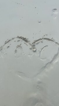    Ocean wave washes over a heart and initials drawn in beach sand.