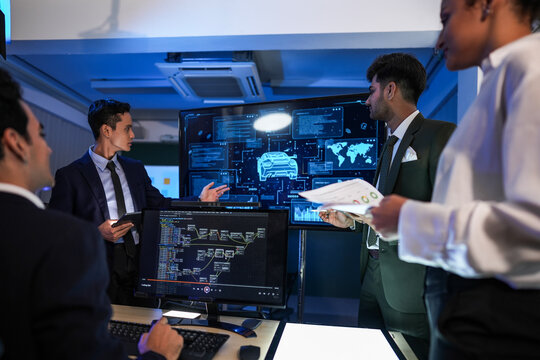 A specialist compares the printed spec against the screen with the car rendered in blue wireframe on the display behind and the pages change hands again in a dark modern office