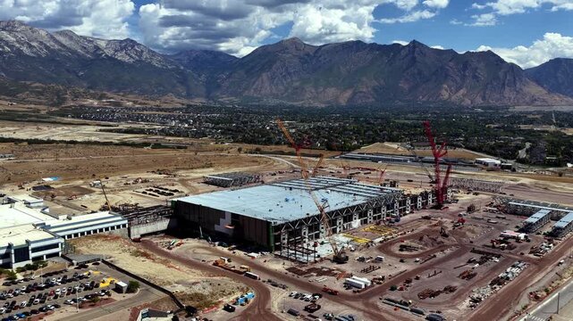 Texas Instruments LFAB2 semiconductor fab under construction beside LFAB1, with cranes, construction yards and support buildings against the Wasatch Range in Lehi, Utah, USA, drone pullback