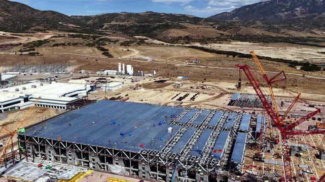 Texas Instruments LFAB2 300mm semiconductor wafer fab under construction, with steel framing, tower cranes, industrial buildings and Wasatch Mountain foothills at Lehi, Utah, USA, drone lateral slide