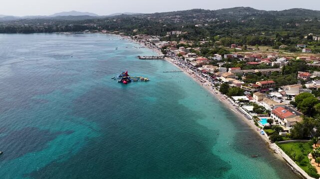 Aerial drone view of beautiful Ipsos beach coastline in Corfu Greece with clear turquoise sea water resort town villas green hills and floating inflatable park