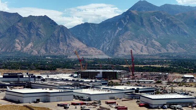 Texas Instruments LFAB2 construction beside the LFAB1 semiconductor fab campus, with cranes, industrial buildings and the Wasatch Range in Lehi, Utah, USA, drone forward flyover