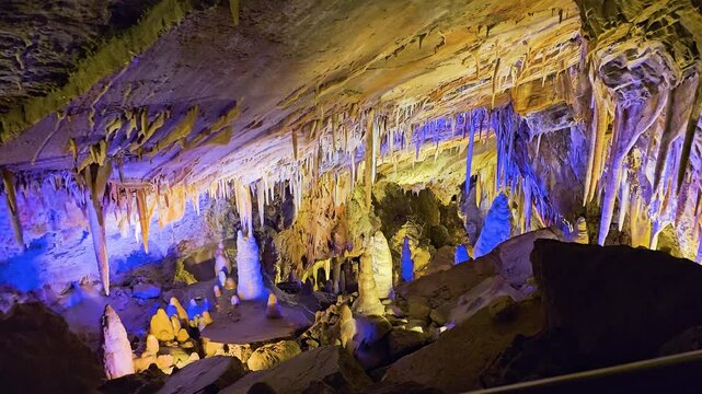 Colorfully illuminated limestone cave filled with stalactites, stalagmites, mineral formations, rugged rock walls, and dramatic blue and amber lighting inside a vast underground cavern.