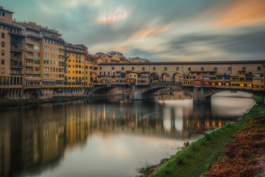 View of the historic Ponte Vecchio bridge spanning the Arno River with medieval architecture, colorful buildings, and reflections on the water in Florence, Tuscany, Italy.
