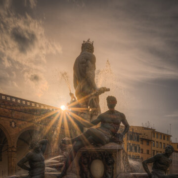 View of the Fountain of Neptune in Piazza della Signoria at sunset with a starburst effect behind the marble statue and historic buildings in Florence, Tuscany, Italy.