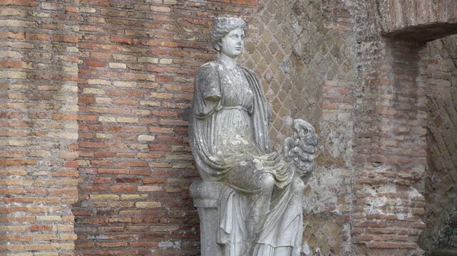 Seated classical female statue holding a broken cornucopia leans against an old masonry backdrop in Ostia Antica.