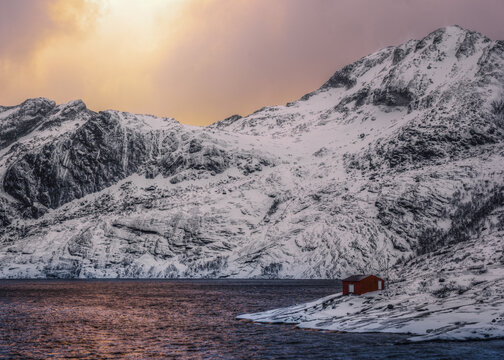 View of a red cabin on a snowy shore by a dark fjord with towering snow-covered mountains in the background under a golden sky in Nordland, Norway.