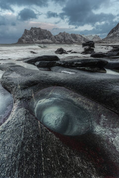 View of a smooth circular rock pool in the foreground with dark wet rocks, a sandy beach, and snow-capped mountains under a dramatic cloudy sky in Lofoten, Nordland, Norway.