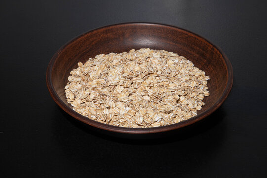 Rolled oats flakes background closeup with raw oat flakes texture, organic healthy fitness breakfast cereal pile in rustic clay bowl over dark surface, horizontal copy space for packaging design.