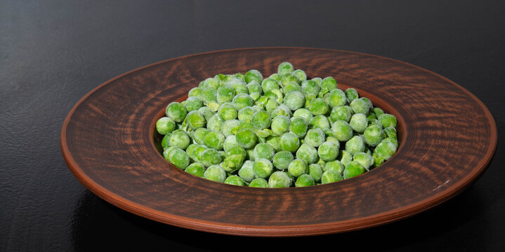 Frozen green peas background closeup with ice crystals and frost texture, organic sweet pea seeds on rustic clay plate over dark surface, horizontal top view copy space for packaging design.