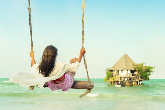 Young pretty woman tourist in a pink sarong relaxes on a beach swing overlooking the iconic Rock Restaurant in Zanzibar. Peaceful coastal scene with turquoise water and blue sky