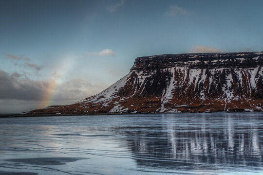 View of a snow-capped mountain rising above a frozen lake with a faint rainbow in the sky in Iceland.