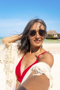 Happy mid adult caucasian woman in red bikini sunglasses and white lace beach sarong shawl taking selfie portrait on tropical sandy beach clear blue sky shore Zanzibar Tanzania