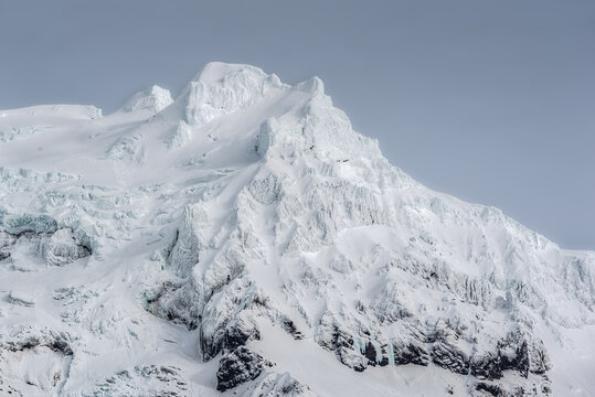 View of a rugged, snow-covered mountain peak with deep glacial ice textures and blue-tinted shadows under an overcast sky in Iceland.