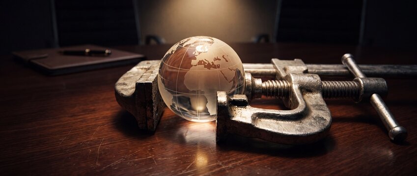 Glass globe held in metal vise grip on dark wooden table representing environmental pressure and global crisis in executive boardroom.