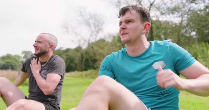 Two men resting on grass after jogging in park, enjoying sunny day