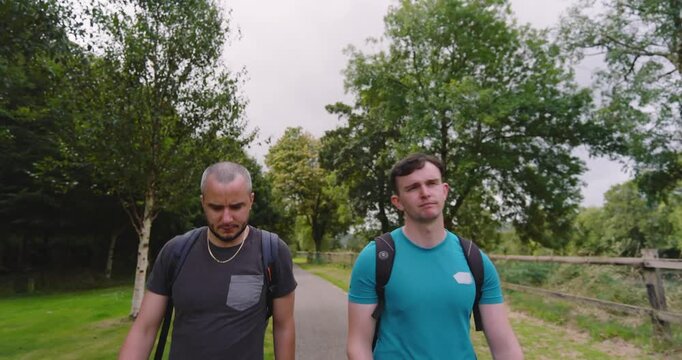 Two men walking in park with backpacks, enjoying conversation and nature. There is a lot of trees an