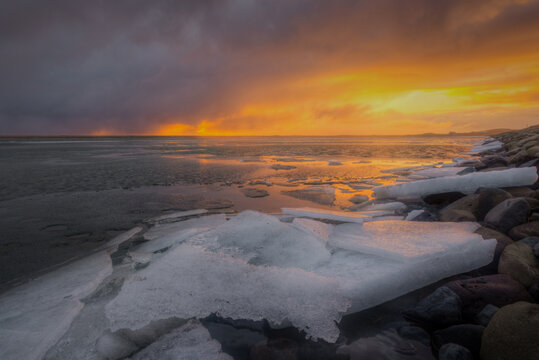 View of ice chunks on a rocky shoreline during a vibrant sunset with dramatic clouds over the water in Iceland.