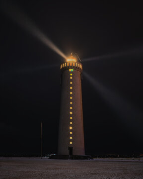 View of the illuminated Grotta Lighthouse casting a bright beam into the dark night sky over the snowy landscape in Iceland.