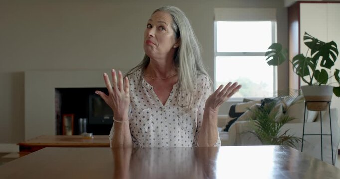 Mature senior woman at home expressing surprise with hands raised, sitting at table